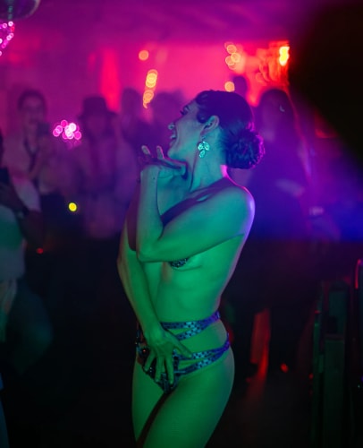 Personne dansant dans une ambiance de boîte de nuit avec des lumières colorées violettes et vertes, portant des accessoires et une tenue de scène minimaliste, entourée d'un public flou en arrière-plan.