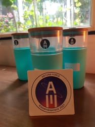 Trois verres en plastique transparent avec liquide bleu clair, posés sur une table en bois, avec un autocollant devant portant le texte « ARTLINE FORCE Rassemblement 2021 » et un logo circulaire. En arrière-plan, une fenêtre avec des plantes visibles à travers.