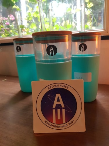 Trois verres en plastique transparent avec liquide bleu clair, posés sur une table en bois, avec un autocollant devant portant le texte « ARTLINE FORCE Rassemblement 2021 » et un logo circulaire. En arrière-plan, une fenêtre avec des plantes visibles à travers.