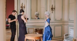 Une femme en costume d'époque bleu avec une perruque blanche posant devant un grand miroir dans une pièce élégante, pendant qu'un homme et une femme discutent à côté d'elle, la femme tenant un appareil photo.