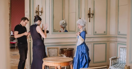 Une femme en costume d'époque bleu avec une perruque blanche posant devant un grand miroir dans une pièce élégante, pendant qu'un homme et une femme discutent à côté d'elle, la femme tenant un appareil photo.