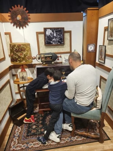 Un homme assis sur une chaise et deux enfants penchés sur un bureau dans une pièce décorée vintage avec une lampe à motifs floraux, un lampadaire en forme de soleil et plusieurs cadres accrochés aux murs.