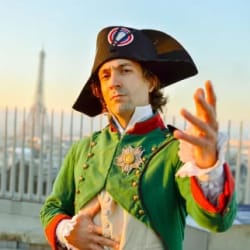 Un homme habillé en costume historique vert et noir avec une coiffe tricorne, faisant un geste avec la main, avec la Tour Eiffel floue en arrière-plan au coucher du soleil.