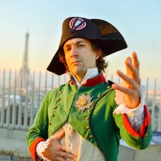 Un homme portant un costume historique de Napoléon, avec un chapeau bicorne noir orné d'un symbole, pose avec un bras tendu devant un paysage urbain au coucher du soleil.