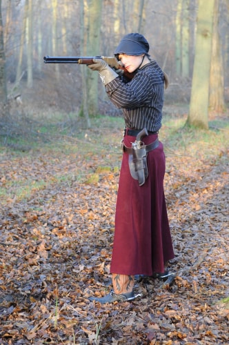 Femme habillée en tenue d'époque avec chapeau noir, gants et jupe longue bordeaux, visant avec un fusil dans une forêt en automne, des feuilles mortes au sol.