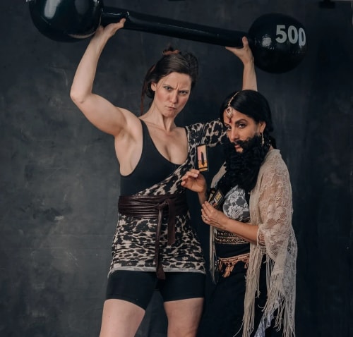 Une femme musclée soulève une barre de poids noire marquée 500 au-dessus de sa tête, à côté d'une autre femme déguisée avec une fausse barbe noire, portant un châle en dentelle et tenant des cartes entre ses mains, sur un fond sombre.