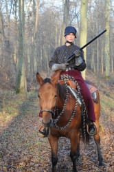 Personne en tenue d'époque, coiffée d'un bonnet noir, montant un cheval marron dans une forêt en automne, tenant un fusil.