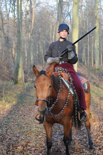 Personne en tenue d'époque, coiffée d'un bonnet noir, montant un cheval marron dans une forêt en automne, tenant un fusil.