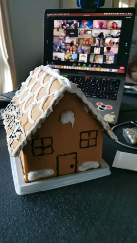 Une maison en pain d'épice décorée de glaçage blanc devant un ordinateur portable affichant une réunion en visioconférence avec plusieurs personnes.