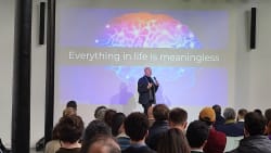 Un homme donne une présentation devant un auditoire avec une image d'un cerveau coloré projetée en arrière-plan et le texte en anglais : "Everything in life is meaningless".