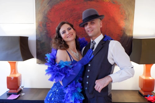 Un couple habillé en tenue de soirée vintage pose joyeusement devant un mur avec une grande peinture abstraite rouge. La femme porte une robe bleue avec un boa à plumes assorti et des gants, tandis que l'homme porte un gilet, une chemise blanche, une cravate et un chapeau.