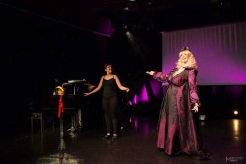 Deux femmes sur scène, l'une en robe longue violette et perruque blonde claire, l'autre en tenue noire près d'un piano avec un panier de fruits, éclairage violet en arrière-plan.