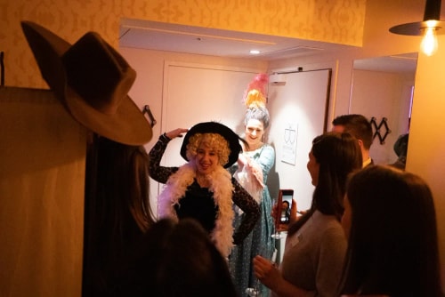 Groupe de personnes dans un couloir, certaines vêtues de costumes avec chapeaux et accessoires, en train de sourire et de prendre des photos dans une ambiance chaleureuse.