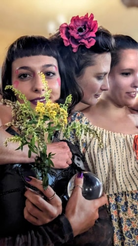 Trois femmes rapprochées, la première tenant un bouquet de fleurs jaunes et une boule en verre, avec un maquillage coloré et une fleur rouge dans les cheveux de la deuxième femme.