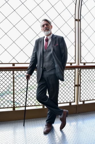 Homme barbu portant un costume gris foncé avec une cravate bordeaux, tenant une canne noire, debout dans un espace avec des grilles métalliques en arrière-plan.
