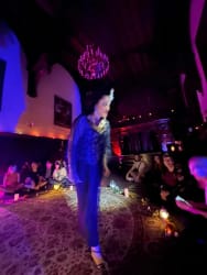 Personne en tenue élégante marchant au centre d'une pièce faiblement éclairée avec des spectateurs assis autour, ambiance tamisée avec lumières violettes et bougies.