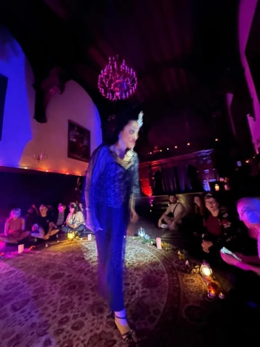 Personne en tenue élégante marchant au centre d'une pièce faiblement éclairée avec des spectateurs assis autour, ambiance tamisée avec lumières violettes et bougies.