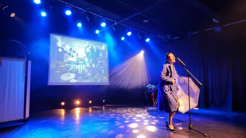 Artiste féminine sur scène sous un éclairage bleu, chantant devant un micro, avec un écran projetant une image en arrière-plan et un paravent blanc sur le côté gauche.