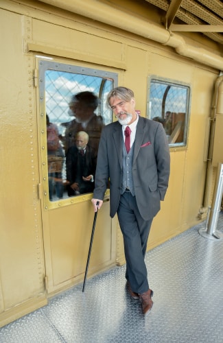 Un homme élégant portant un costume gris foncé avec une cravate rouge, une pochette rouge et une canne noire, se tient en appui contre une porte jaune avec deux petites fenêtres, l'une d'elles reflétant une autre personne assise à l'intérieur.
