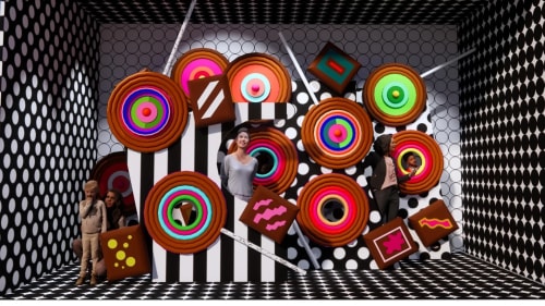 Une pièce décorée avec des motifs géométriques en noir et blanc, contenant de grandes décorations colorées semblant être des bonbons ou des gâteaux, avec plusieurs personnes observant ou interagissant avec ces éléments.
