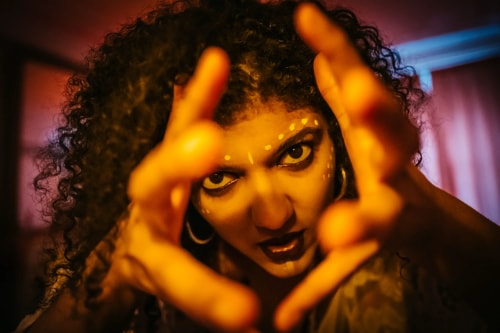 Une personne avec des cheveux bouclés et des peintures faciales blanches regarde intensément vers l'appareil photo, ses mains devant elle formant un cadre.