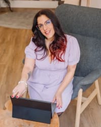 Frances Vieras-Blanc assise dans un fauteuil gris devant une tablette posée sur une table en bois, dans une pièce avec un sol en parquet.