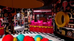 Intérieur coloré avec des murs décorés de motifs abstraits noir rouge jaune, un canapé rouge avec des coussins roses vif, un miroir ovale reflétant une personne debout, un sol en damier noir et blanc et des balles multicolores au premier plan.