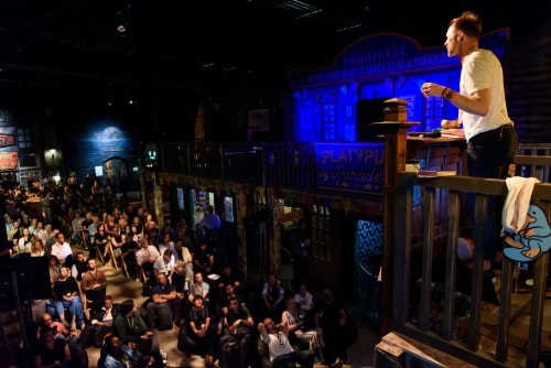 Un orateur debout sur une scène en bois parle devant un public nombreux assis dans une salle avec un éclairage tamisé et des décors rappelant un village ou une fête foraine.