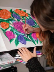 Une femme aux longs cheveux peint avec un pinceau une grande composition de fleurs colorées orange, roses et violettes aux feuilles vertes, sur une feuille posée devant elle.