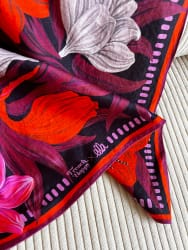 Foulard en soie à motif floral de grandes fleurs rouges, roses et grises sur fond sombre, bordé d'un liseré orange et de pois roses, posé en diagonale sur un tissu côtelé beige.