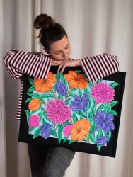 Une femme aux cheveux attachés, vêtue d'un haut rayé, tient contre elle une peinture encadrée de noir représentant des fleurs colorées : orange, violettes et roses, sur un fond bleu clair feuillu.