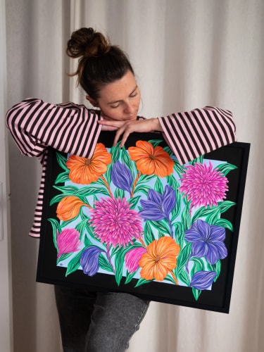 Une femme aux cheveux attachés, vêtue d'un haut rayé, tient contre elle une peinture encadrée de noir représentant des fleurs colorées : orange, violettes et roses, sur un fond bleu clair feuillu.