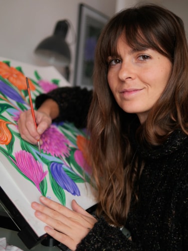 Une femme aux cheveux bruns, vêtue d'un pull noir, peint des fleurs colorées sur une toile blanche posée sur un chevalet, en regardant vers l'objectif avec un sourire doux.