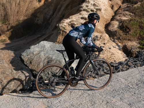 Une cycliste équipée d'un casque et d'une tenue de sport fait du vélo de course sur un chemin rocailleux en extérieur.