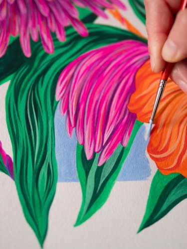 Une main utilise un pinceau fin pour peindre une illustration colorée de fleurs aux pétales roses et orange vif entourées de feuilles vertes sur un fond blanc avec un carré bleu clair.