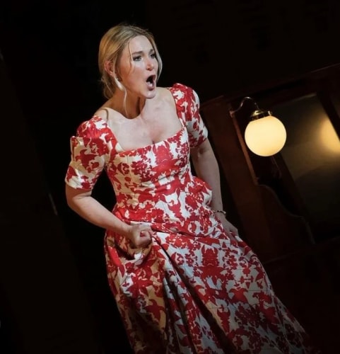 Une femme blonde en robe longue à motifs rouges et blancs, le visage expressif et la bouche ouverte, semble interpréter une scène dramatique ou chanter sur une scène de théâtre peu éclairée.