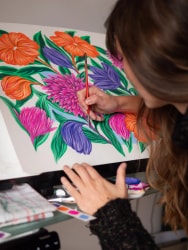 Une artiste aux longs cheveux bruns peint une illustration florale aux couleurs vives avec un pinceau fin sur un chevalet.