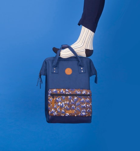 Un pied en chaussette rayée appuie sur un sac à dos bleu marine avec une poche frontale à motifs floraux sur fond bleu.