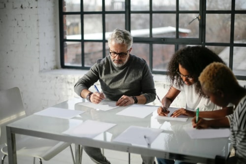 Un homme d'âge mûr avec des lunettes et deux jeunes femmes travaillent ensemble autour d'une table blanche, écrivant sur des feuilles de papier dans un bureau lumineux.