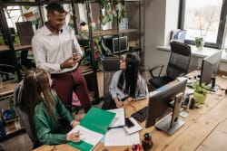 Un homme en chemise blanche discute avec deux femmes travaillant à leur bureau dans un espace de travail collaboratif lumineux.