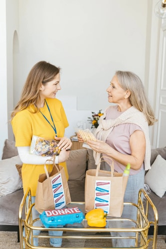 Une jeune bénévole en t-shirt jaune discute et donne des produits à une femme âgée dans un salon, avec des sacs sur un chariot.