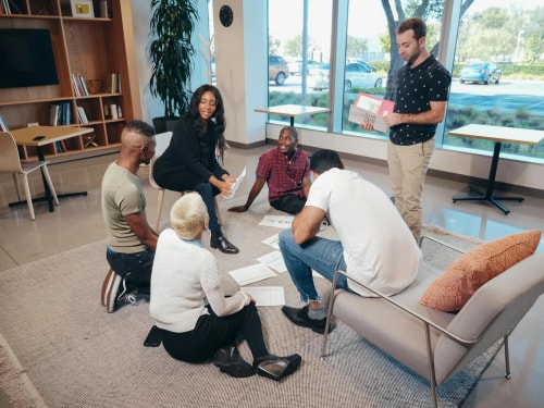 Un groupe diversifié de jeunes professionnels est assis sur un tapis dans un bureau moderne, discutant de documents et de graphiques, avec un homme debout tenant une présentation.