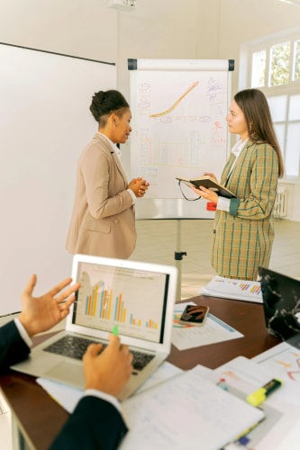 Deux femmes d'affaires discutent devant un tableau blanc avec des graphiques, tandis qu'au premier plan, une personne consulte des données sur un ordinateur portable lors d'une réunion professionnelle.
