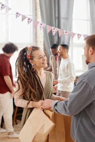 Une femme portant des dreadlocks et un homme se tiennent face à face en échangeant des sacs en papier dans une pièce décorée de guirlandes, avec d'autres personnes en arrière-plan.