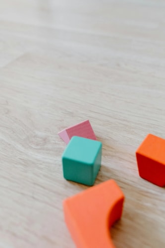 Des blocs de construction en bois colorés, incluant un cube bleu, une forme rose et des pièces orange, sont dispersés sur un sol en bois clair.