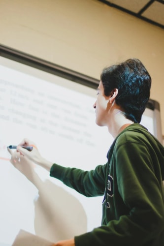 Une personne en sweat-shirt vert écrit sur un tableau blanc interactif dans une salle de classe.