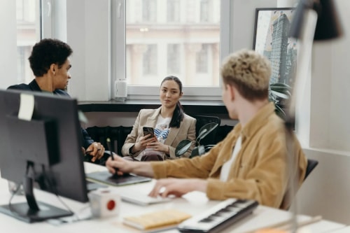 Trois jeunes collègues travaillent ensemble dans un bureau moderne. L'un utilise une tablette graphique, une femme consulte son téléphone et un autre homme est assis en face d'eux.