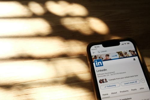 Smartphone affichant la page LinkedIn de développement logiciel avec 24 millions d'abonnés, posé sur une surface en bois claire avec des ombres douces