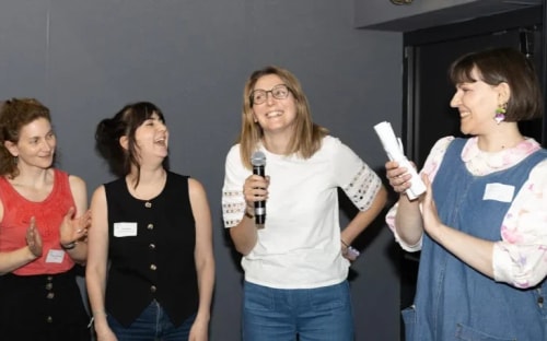 Quatre femmes souriantes se tiennent devant un mur gris, l'une tenant un micro tandis qu'une autre tient un papier. Les trois autres applaudissent lors d'une présentation ou d'un événement.