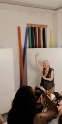 Une femme âgée souriante pose avec élégance devant des rouleaux de papier de fond dans un studio, photographiée par plusieurs personnes au premier plan.